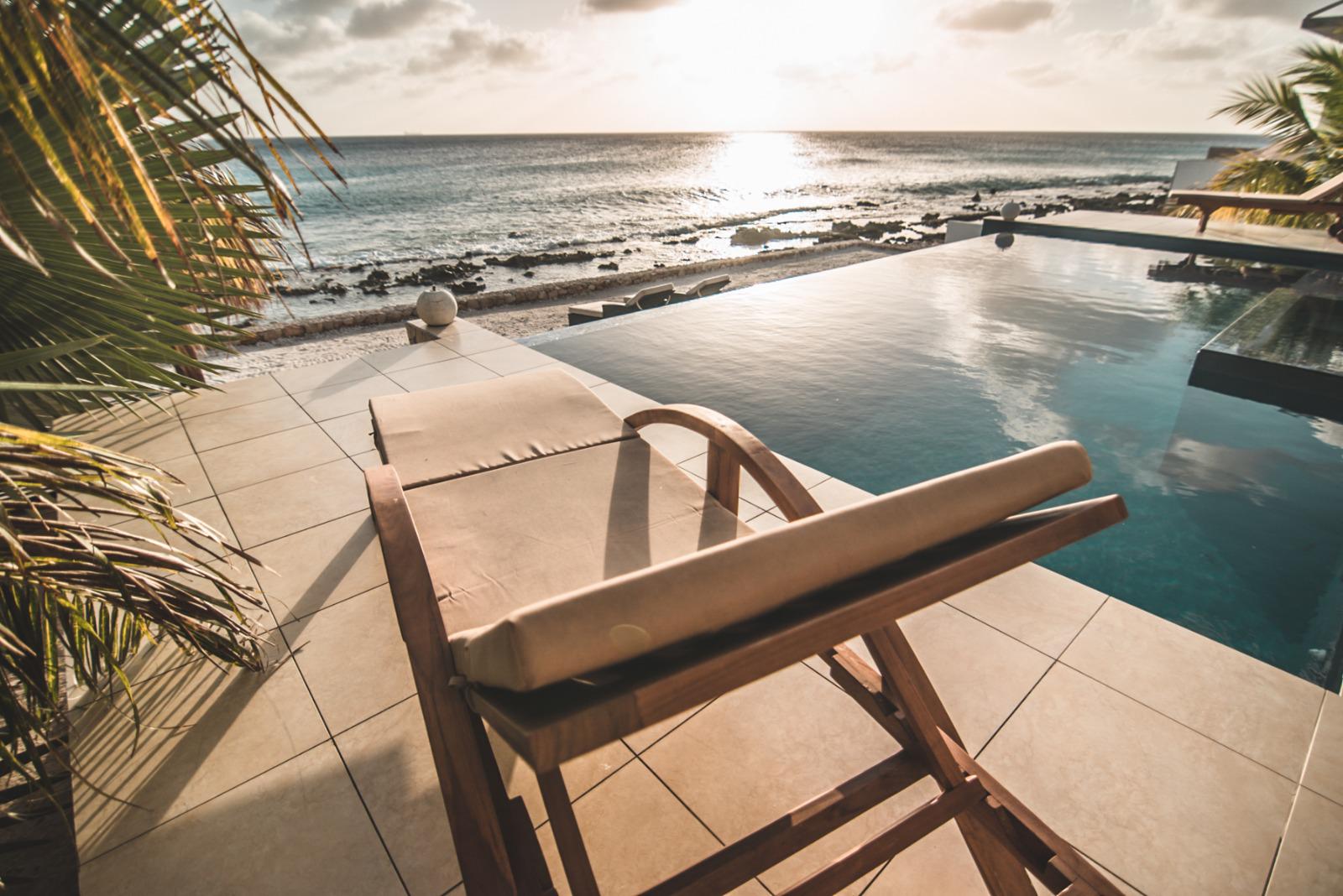 Luxury Bonaire villa with an oceanfront infinity pool and teak lounge chair overlooking the Caribbean Sea at sunset.